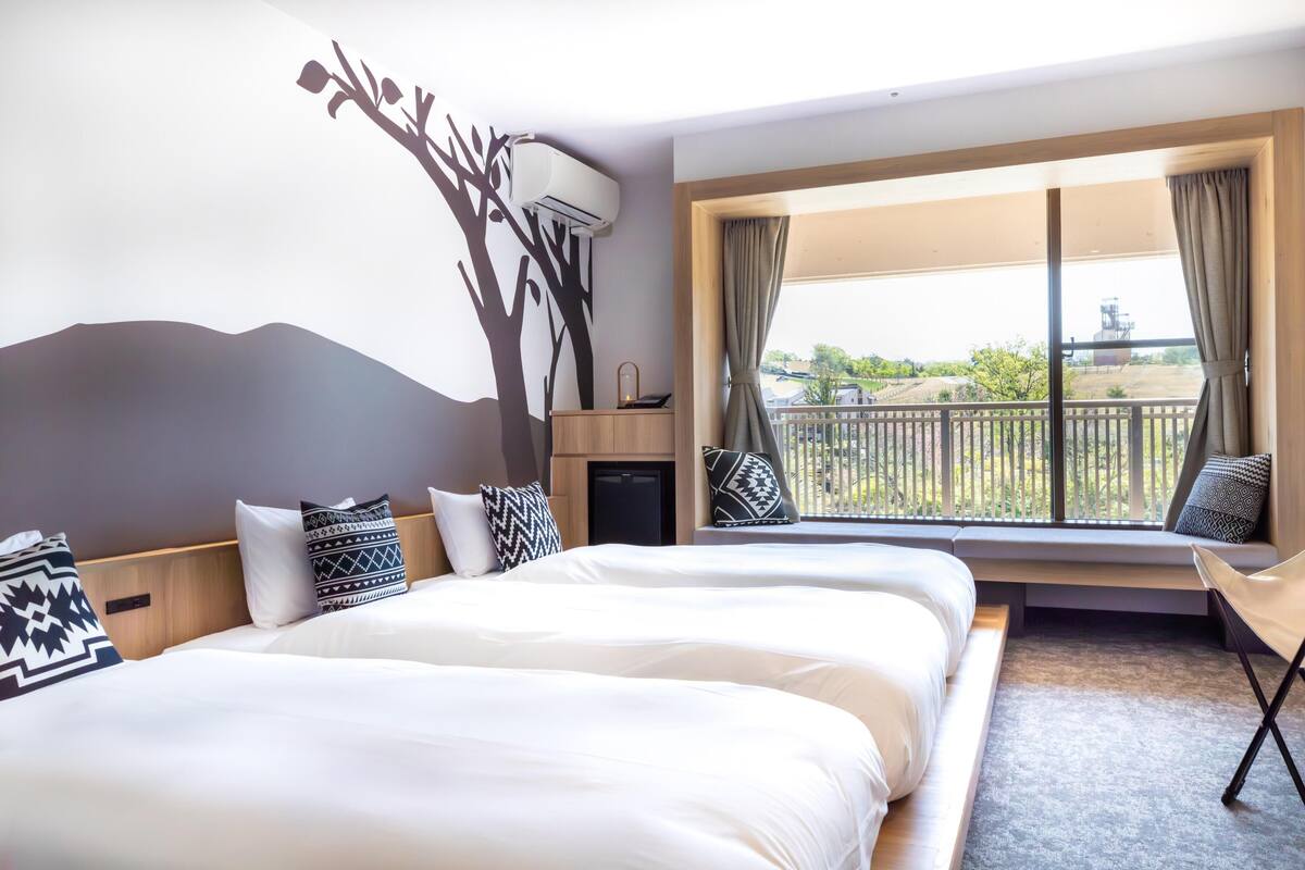A hotel room with two beds, a large window with curtains, a wall-mounted air conditioner, and a view of the outdoors.