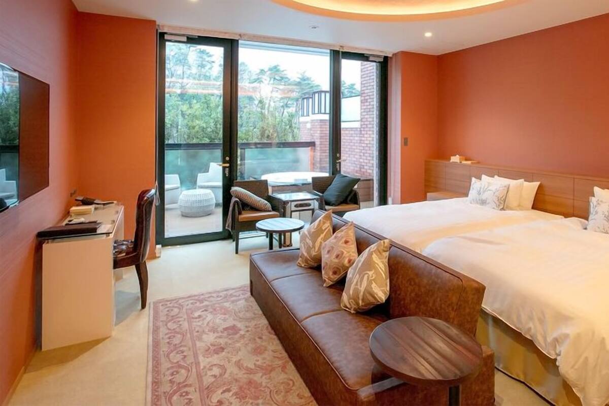 A hotel room with a large bed, a sofa, a desk, and a balcony with outdoor seating.