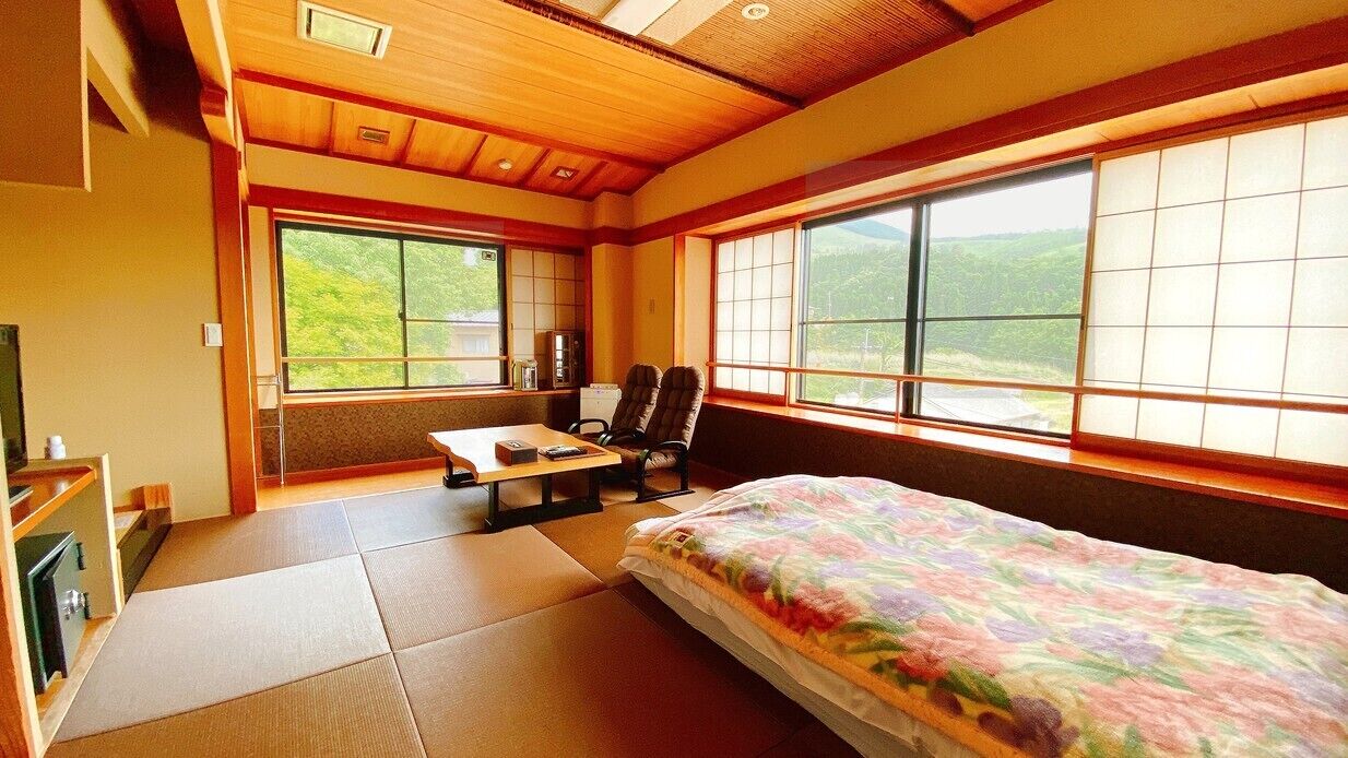 Habitació amb sòls de tatami, un llit amb edredó floral, una taula amb cadira i grans finestres que donen vista a vegetació.