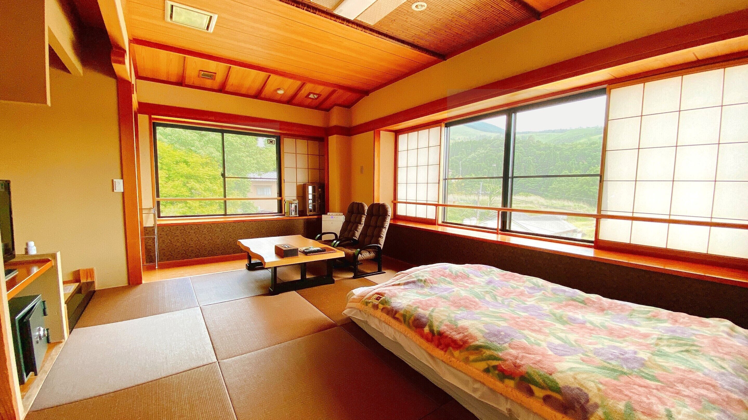 Ein traditioneller japanischer Raum mit Tatami-Boden, einem Bett mit floralem Überwurf, einem Schreibtisch mit Stuhl und großen Fenstern mit Blick auf Grünflächen.