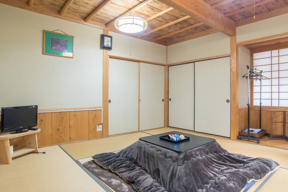 A traditional Japanese room with tatami flooring, a television on a low stand, a futon, sliding doors, and a view through a shoji screen.