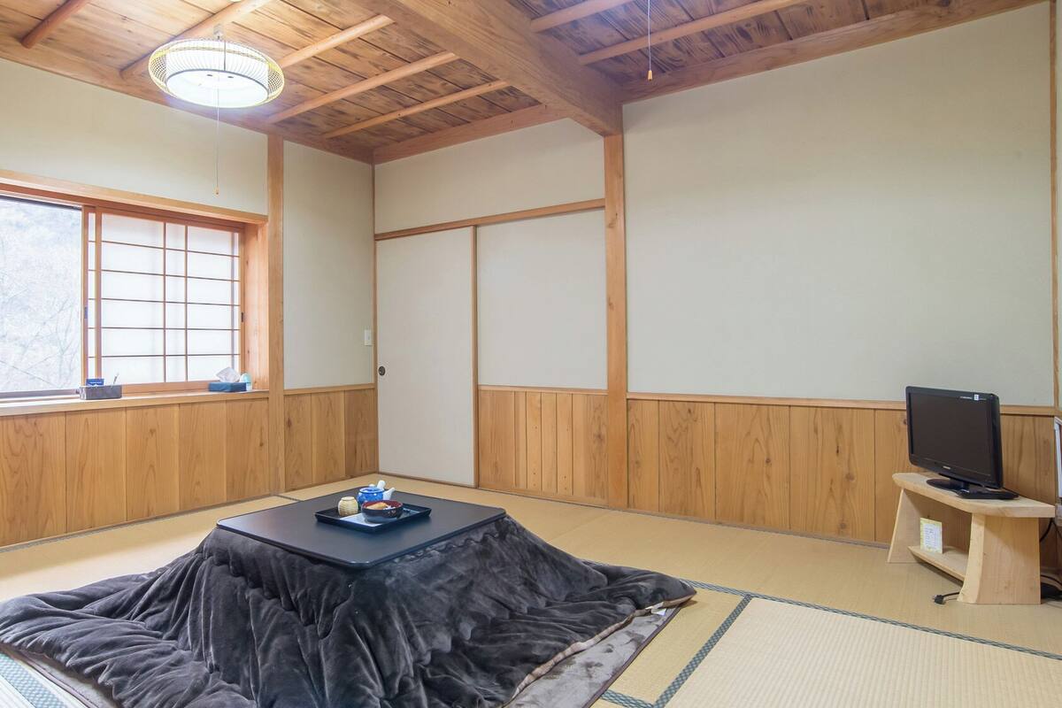 A traditional Japanese room with tatami flooring, a low table with a cushion, a television on a wooden stand, and a shoji screen door.