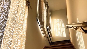Uma escada de madeira com corrimão, cortinas estampadas e luz do sol entrando pela janela.