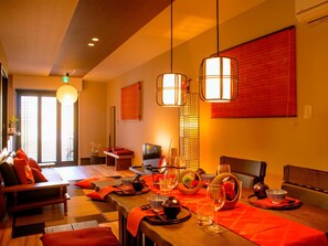 A hotel room with a dining area, a sofa, a TV, and a large window with blinds.