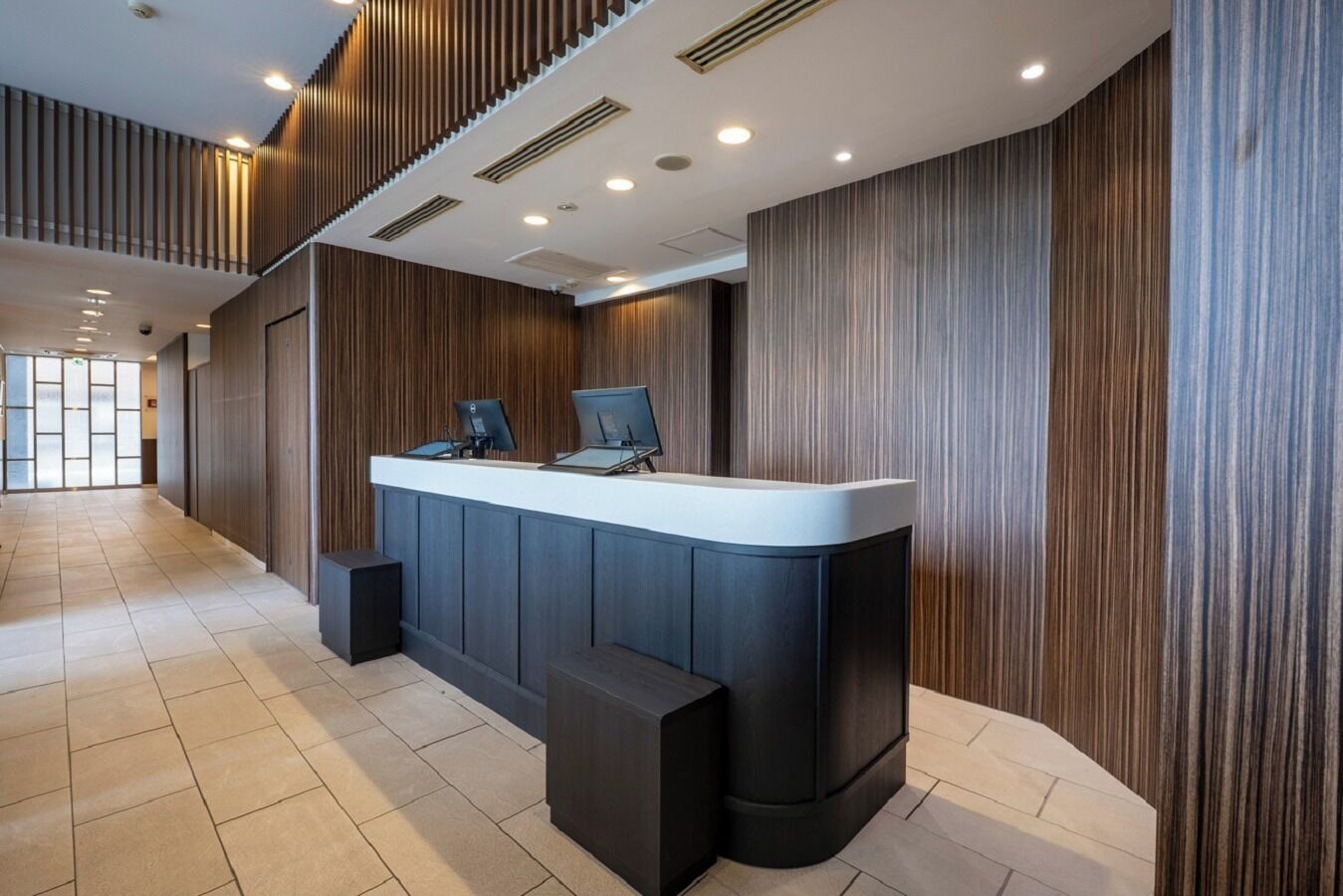 En modern receptionsdisk med två datorskärmar, en platt-tv och en liten svart högtalare. I receptionsområdet finns träpanel på väggarna och ett klinkergolv.