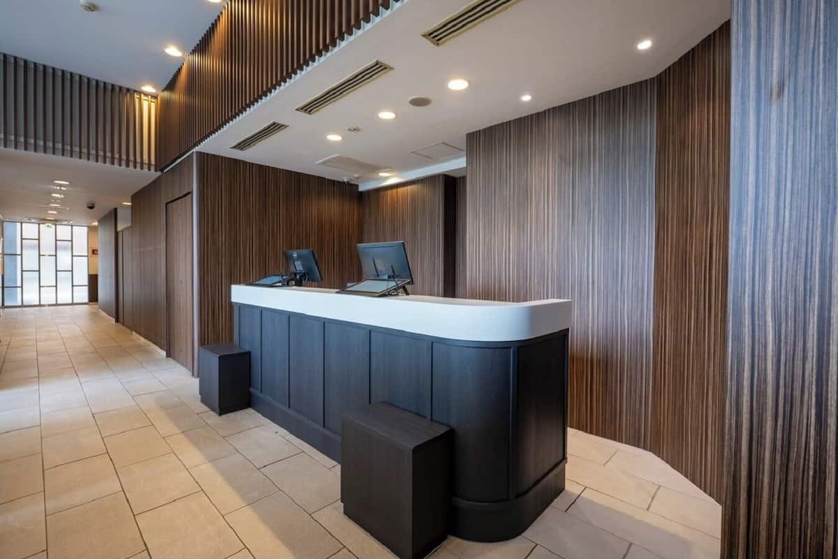Un comptoir d’accueil moderne, doté de deux écrans d’ordinateur, d’un téléviseur à écran plat et d’un petit haut-parleur noir. L’espace d’accueil est agrémenté de panneaux de bois aux murs et d’un sol carrelé.