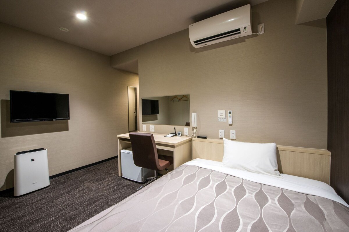A hotel room with a bed, a desk, a chair, a television, an air conditioner, and a mirror.