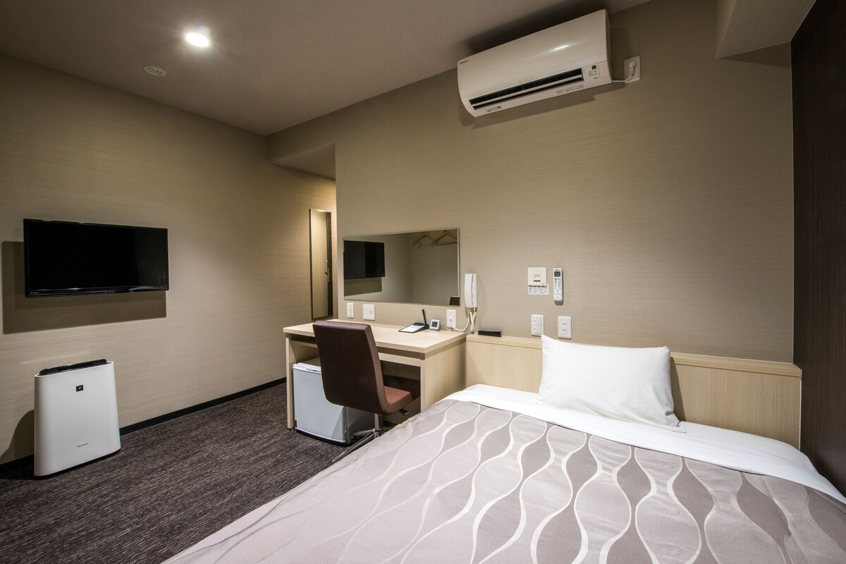 A hotel room with a bed, desk, chair, air conditioning unit, television, and a small refrigerator.