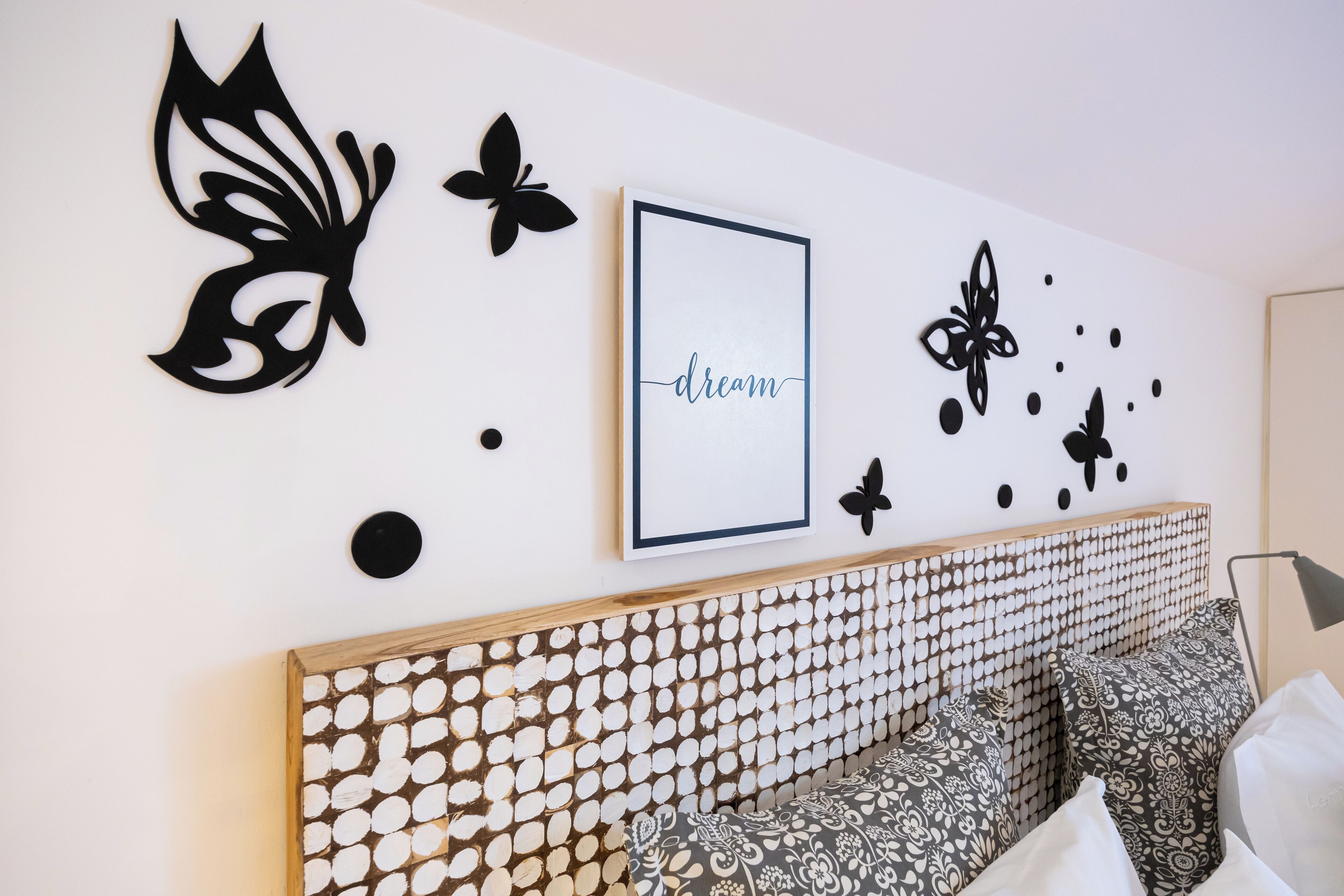 Ein modernes Schlafzimmer mit einem Holz-Kopfteil, gemusterten Kissen und Wandbildern, darunter ein gerahmtes Bild mit dem Wort 'dream' und Schmetterlingsdekorationen.