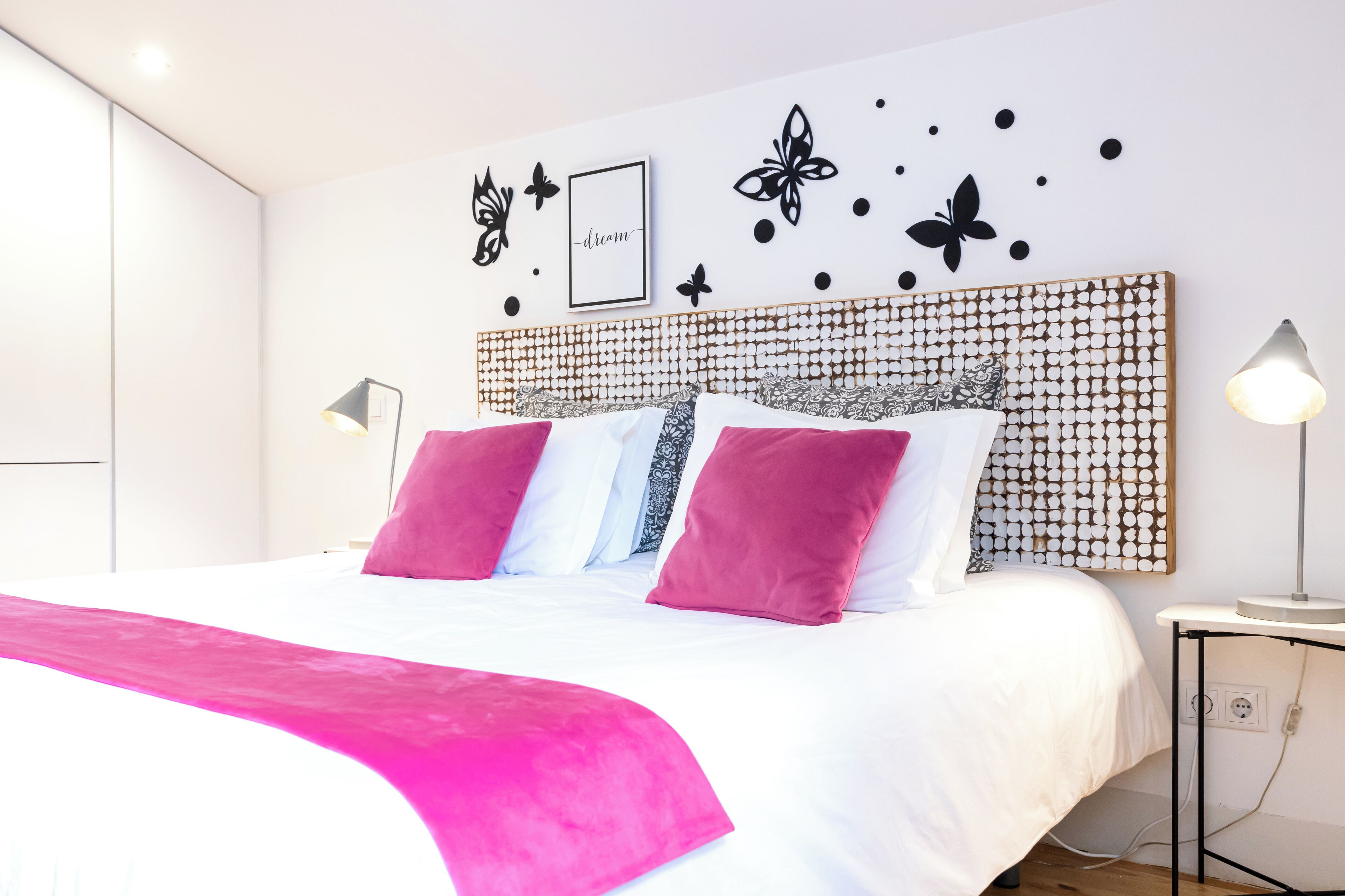 Un lit bien fait, recouvert d’une couvre-lit blanche et rose, avec une tête de lit ornée d’un motif circulaire, et des décorations murales comprenant des papillons et un tableau encadré sur lequel figure le mot « rêve ».