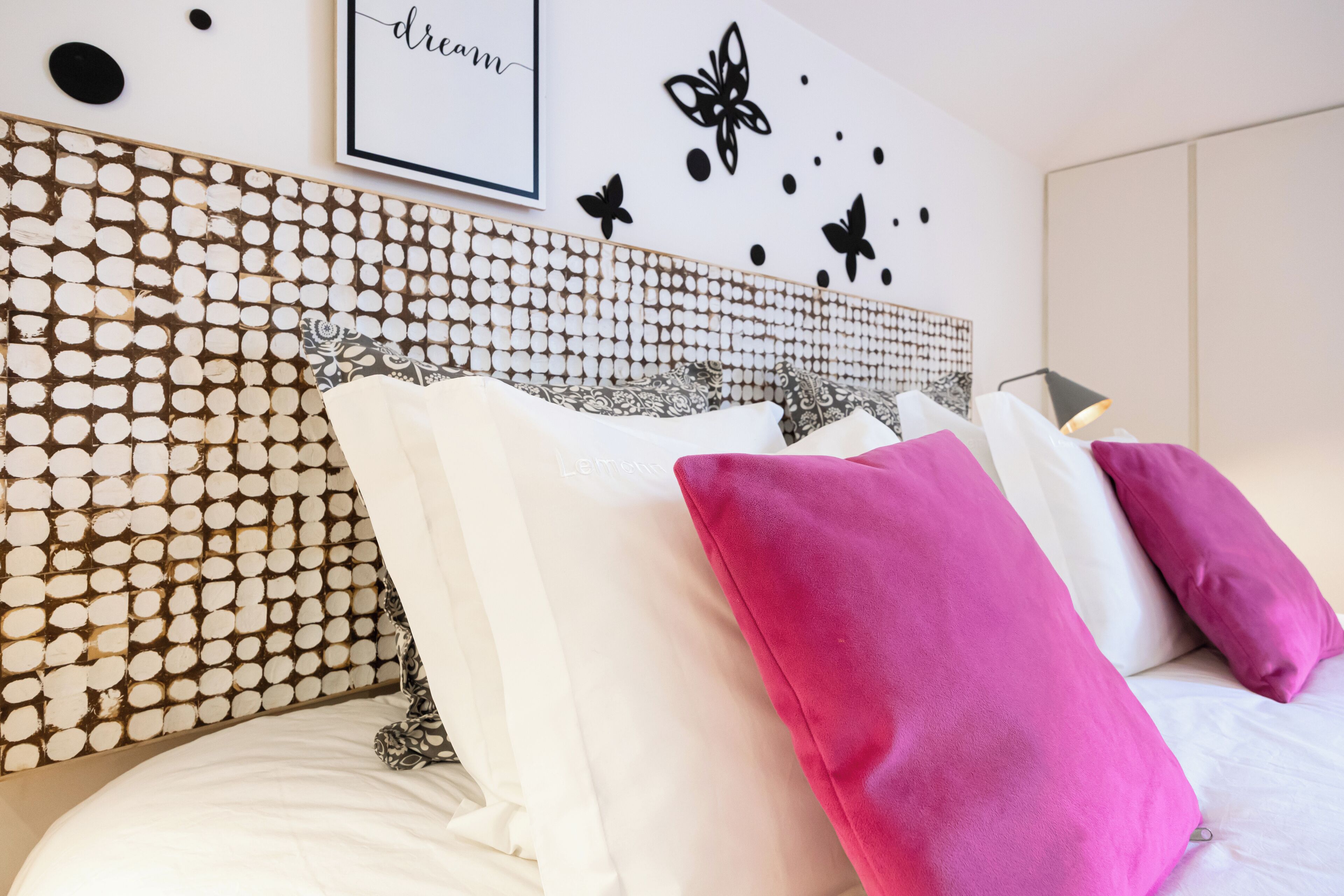 A neatly made bed with white and pink pillows, a decorative headboard with a circular pattern, and a wall with a framed text and butterfly decorations.