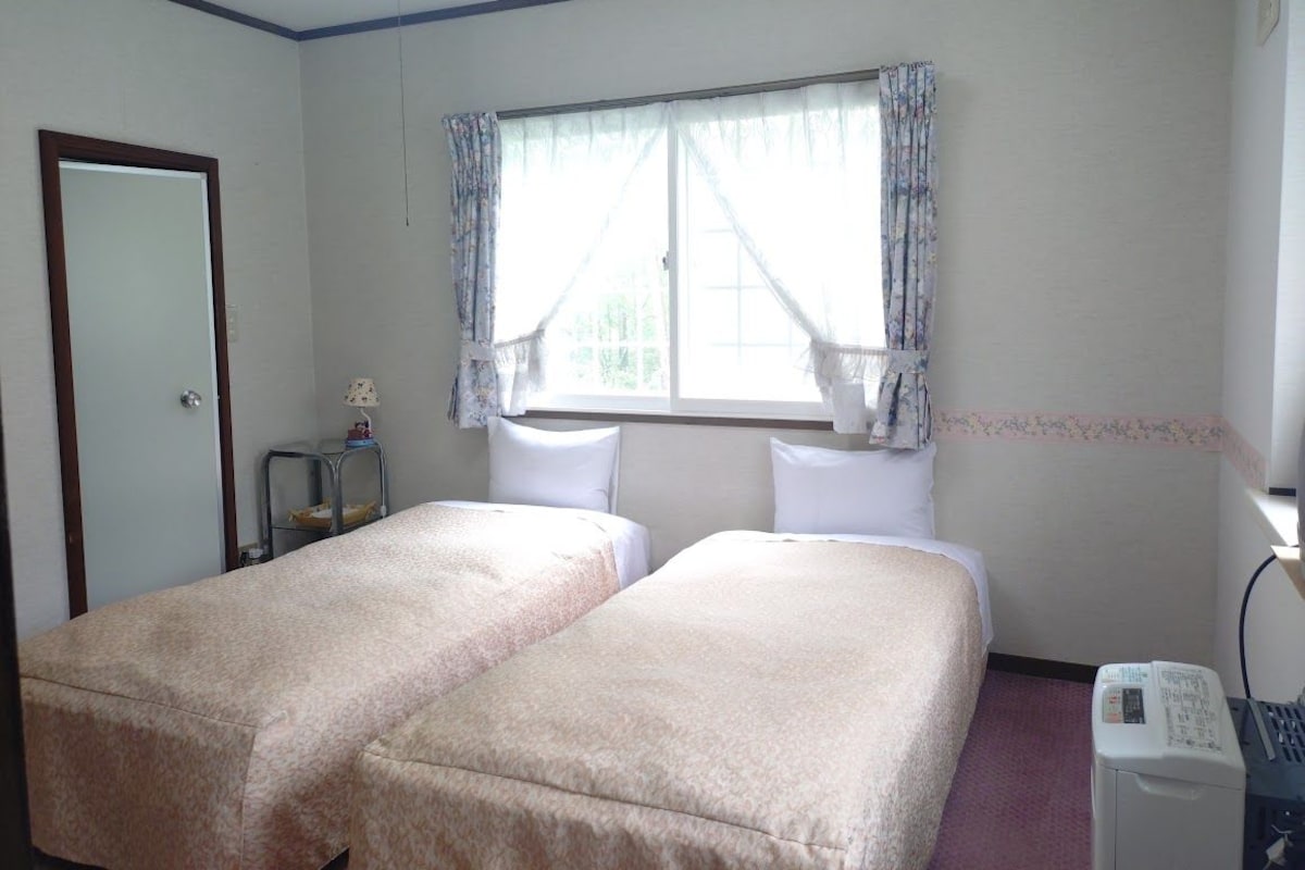 A room with two beds, a window with curtains, a door, a small table with a lamp, and a washing machine.
