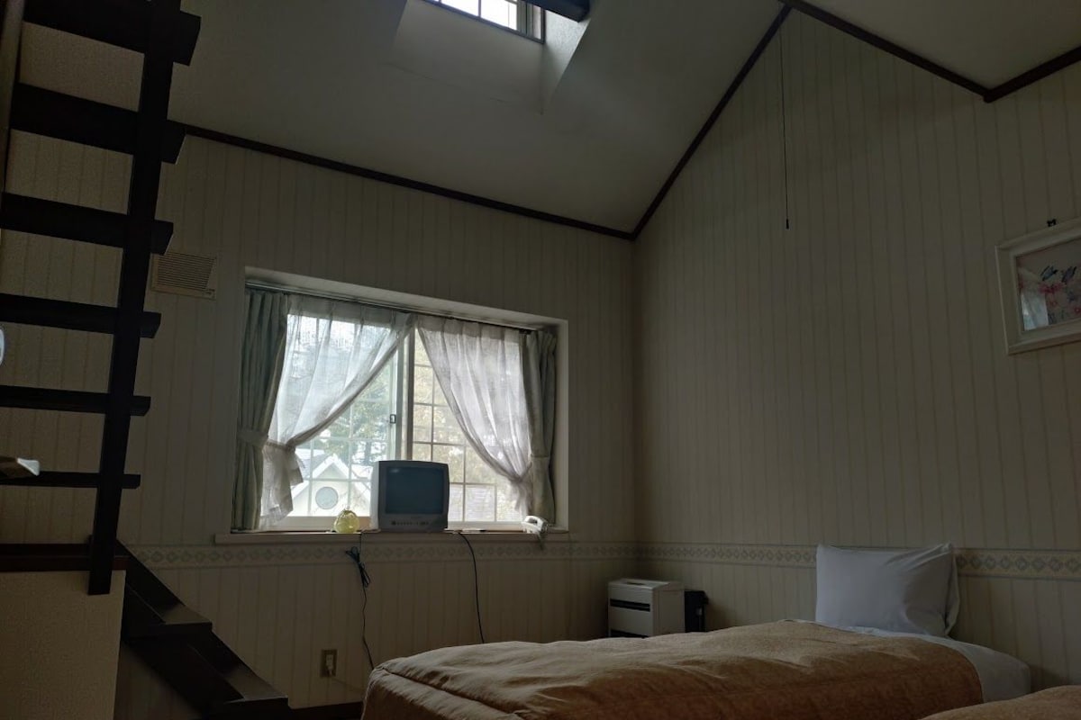 A bedroom with two beds, a window with curtains, a radiator, and a staircase.