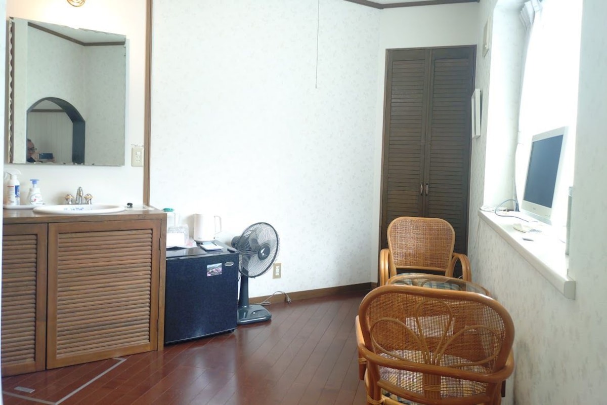 A room with a wooden cabinet, a mirror, a fan, a chair, and a window with a computer monitor.