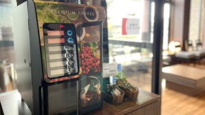 A coffee machine with a digital display and buttons, placed on a wooden counter in a cafe. - Hana Hotel Isesaki (Isesaki)