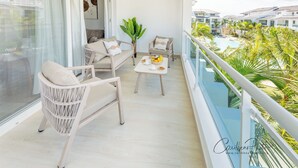 Varanda com mobiliário de vime, mesa de jantar com frutas e vista para uma piscina e edifícios.