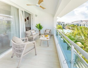 Ein Balkon mit Korbmöbeln, einem Tisch mit Früchten und Blick auf ein Schwimmbad und Gebäude.