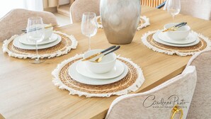 Uma mesa de jantar posta com pratos brancos, talheres dourados e jogos americanos trançados. Um vaso com plumas de capim-dos-pampas está colocado no centro.