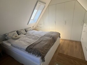 Ein Schlafzimmer mit schrägen Wänden, einem Bett mit gemustertem Überwurf, einem kleinen Dachfenster und weißen Kleiderschränken.
