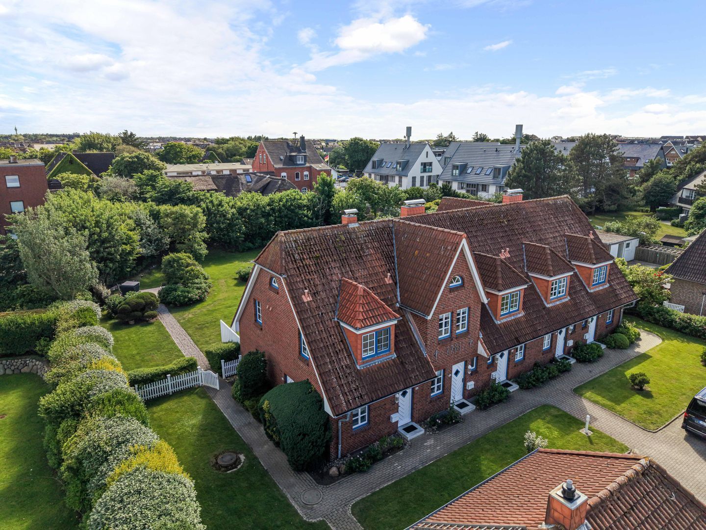 Een groot, twee verdiepingen tellend huis met een rode pannendak, omgeven door verzorgde gazons en een witte hekwerk.