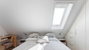 Quarto com teto inclinado, cama com roupa de cama branca, duas mesas de cabeceira, escrivaninha com cadeira e guarda-roupa.