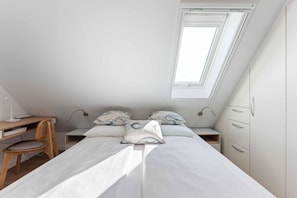 Ein Schlafzimmer mit schrägen Wänden, einem Bett mit weißer Bettwäsche, zwei Nachttischen, einem Schreibtisch mit Stuhl und einem Kleiderschrank.