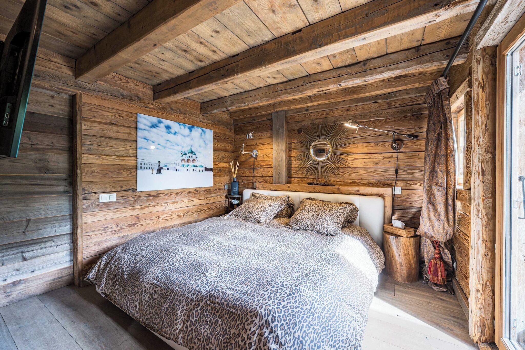 A wooden cabin bedroom with a bed, a wall-mounted picture, and a circular mirror.