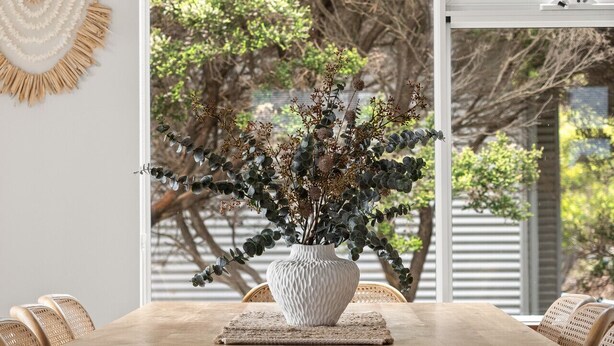 A dining area with a wooden table and woven chairs, a white vase with plants, and a large window with a view of trees. - The Surf Nook | Walk to Beach | Family Friendly (Gracetown)