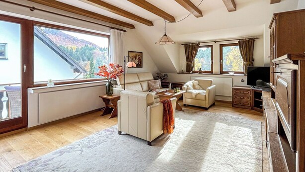 Uma sala de estar acolhedora, com um sofá, poltronas e uma televisão. Há janelas amplas com cortinas, teto de madeira e vista para o exterior.