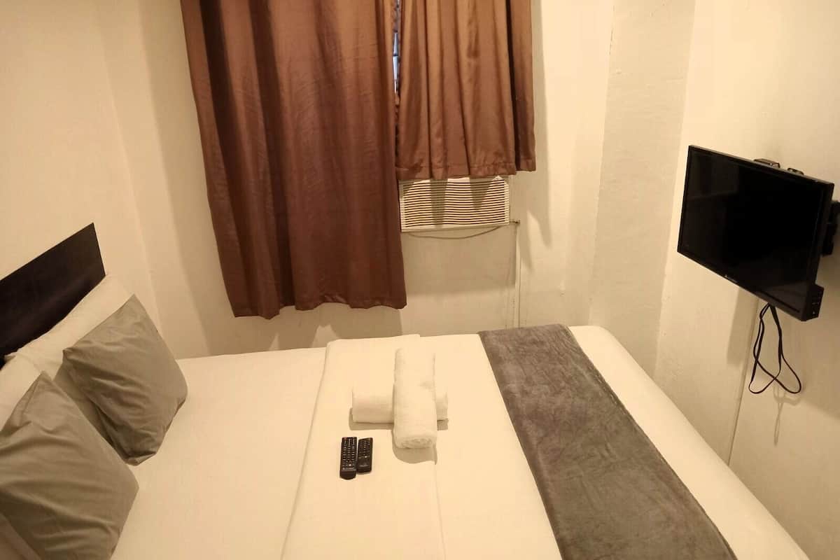 A hotel room with a bed, two pillows, a blanket, a remote control, a TV, and curtains.