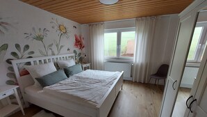 Ein Schlafzimmer mit einem Bett, einem Nachttisch, einem Sessel, einem Heizkörper und einem fenster mit Vorhängen.