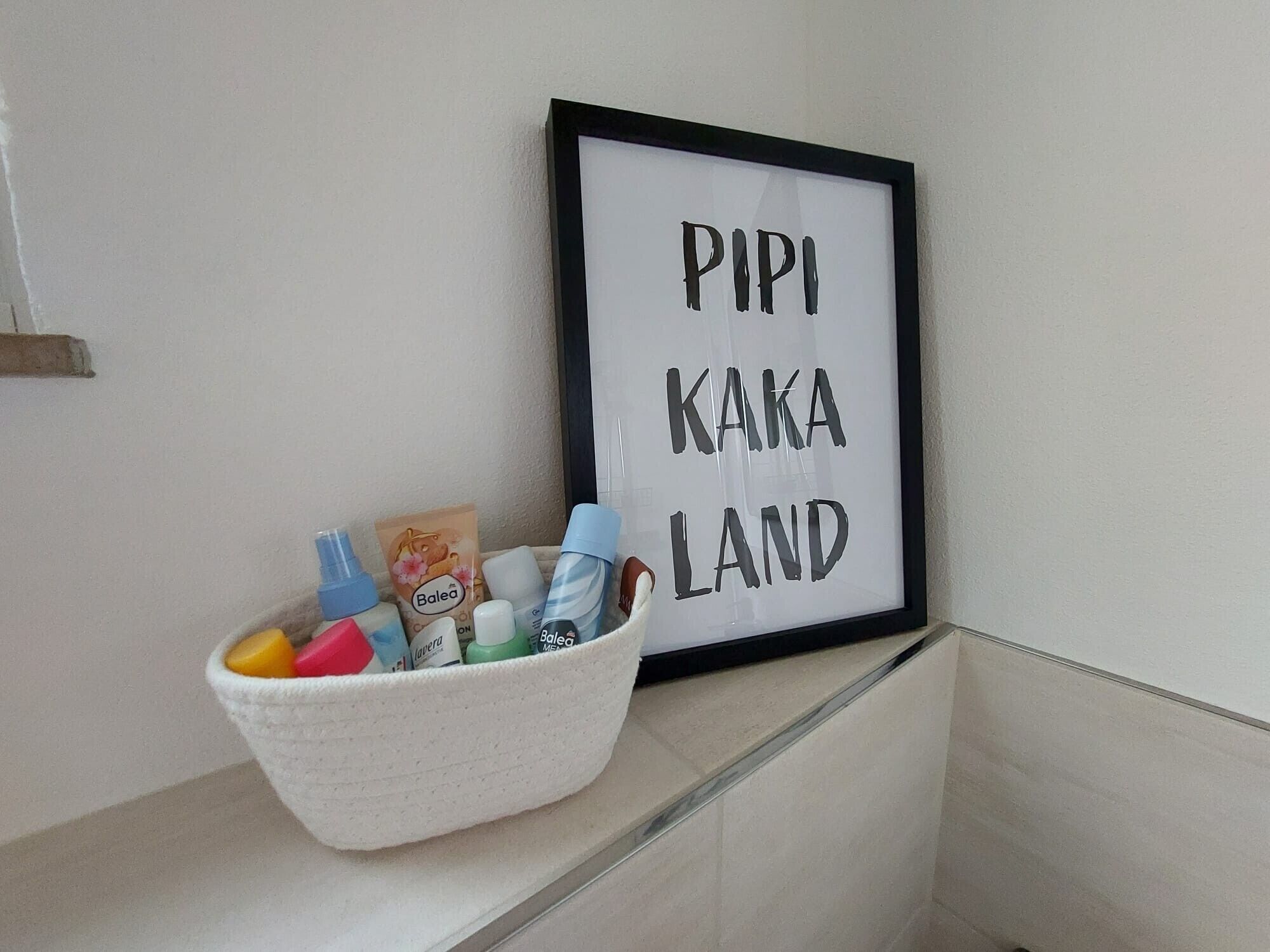 Un letrero en marco con el texto 'PIPI KAKA LAND', una cesta blanca con artículos de aseo y una pared de azulejos.