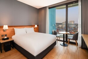 A hotel room with a large bed, two bedside lamps, a small table with a chair, and a view of a cityscape through a large window. - Nova Stay Gangneung (Gangneung)