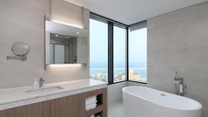 A modern bathroom with a large bathtub, a double sink vanity, a large mirror, and a view of the city and sea through floor-to-ceiling windows. - Nova Stay Gangneung (Gangneung)