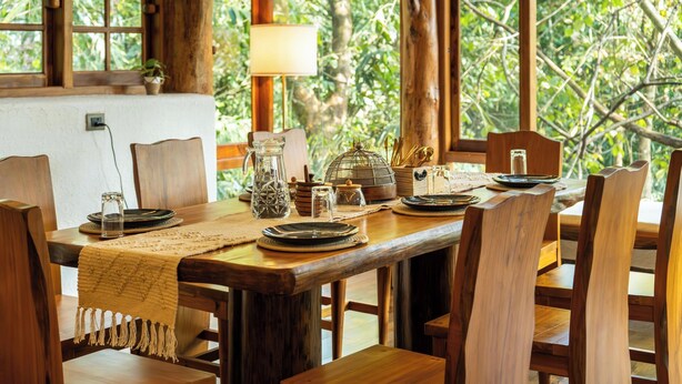 Un tavolo da pranzo in legno apparecchiato con piatti, bicchieri e una brocca, circondato da sedie e con vista su un'abbondante vegetazione attraverso ampie finestre.