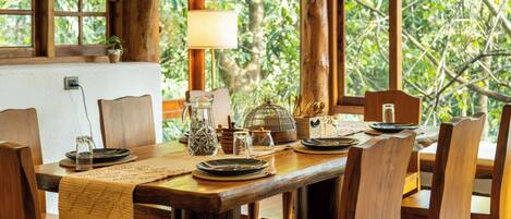 Mesa de madera dispuesta con platos, vasos y una jarrón, rodeada de sillas con vistas a exuberante follaje a través de grandes ventanales.
