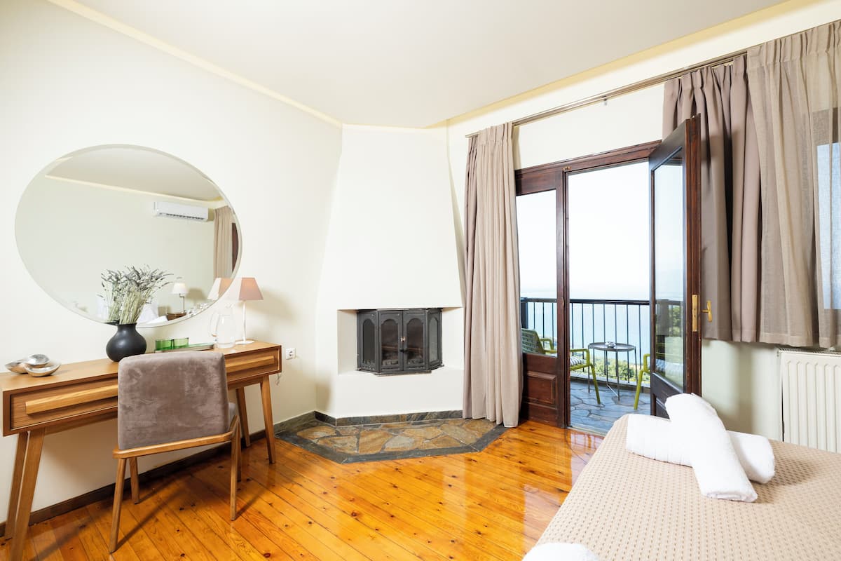 A hotel room with a wooden desk, a chair, a round mirror, a fireplace, a bed with towels, and a balcony with a view.