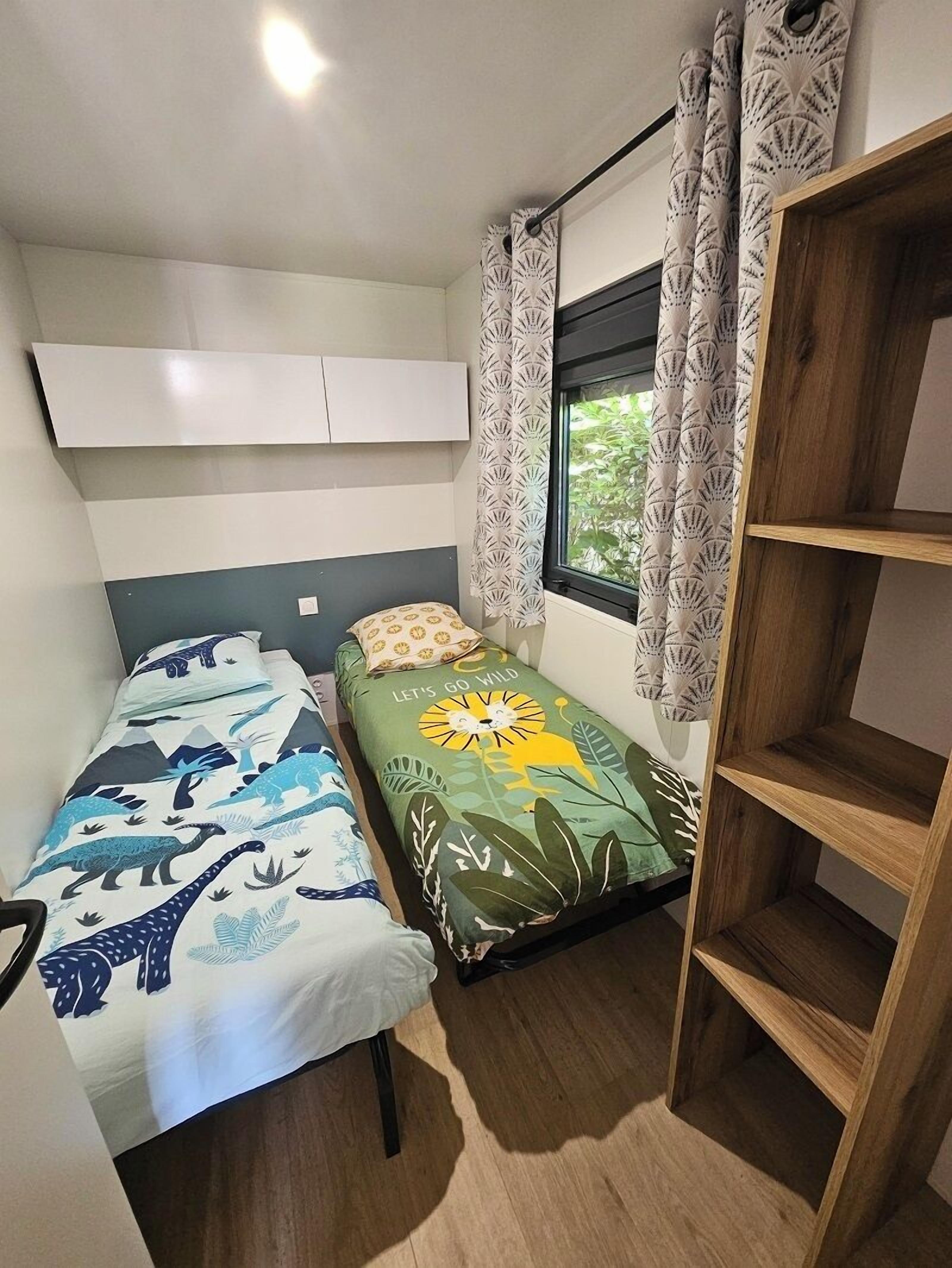 A small, well-lit room with two beds, one with a dinosaur-themed bedspread and the other with a lion and jungle print. There is a window with curtains, a wooden shelf unit, and a white wall-mounted shelf above the beds.