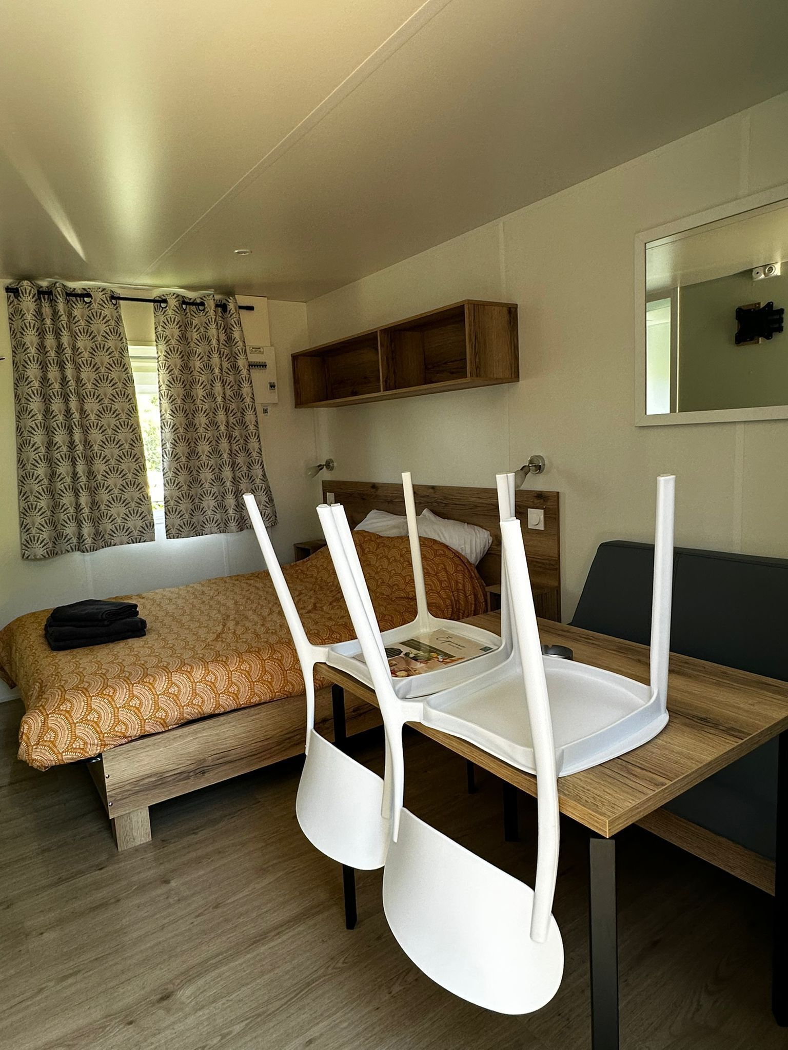 Ein kleines Zimmer mit Bett, Holztisch und Stuhl. Ein Fenster mit Vorhängen, ein Spiegel an der Wand und ein Regal über dem Bett.