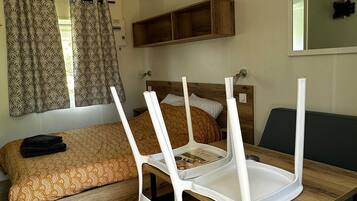 Ein kleines Zimmer mit Bett, Holztisch und Stuhl. Ein Fenster mit Vorhängen, ein Spiegel an der Wand und ein Regal über dem Bett.