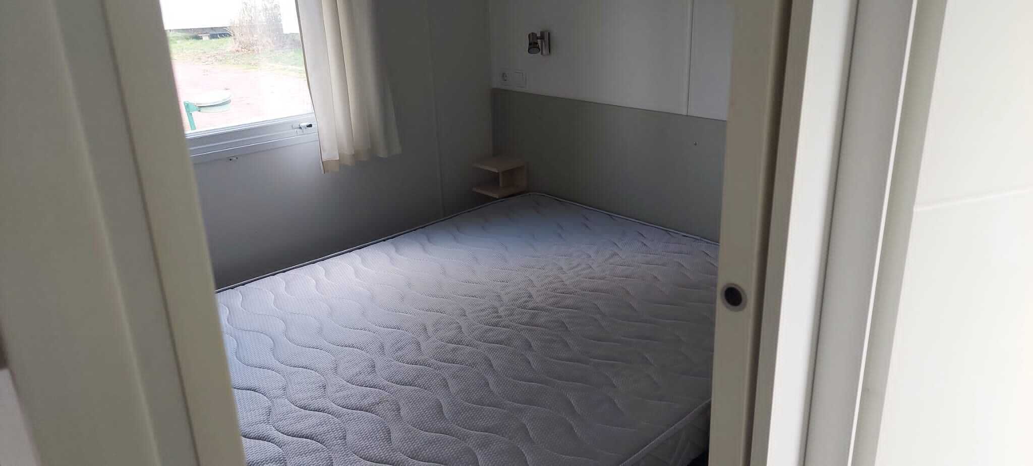 Ein kleiner, schmales Zimmer mit einer Matratze auf dem Boden, einem Fenster mit Vorhängen und einem kleinen Regal an der Wand.