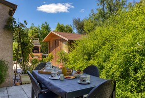 A table set outdoors with breakfast items, surrounded by greenery and a wooden building in the background.