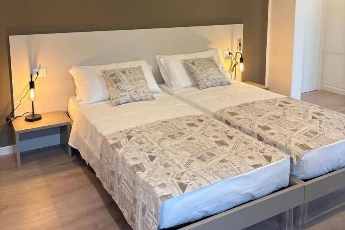 A double bed with a patterned bedspread, two bedside tables with lamps, and a plain wall.