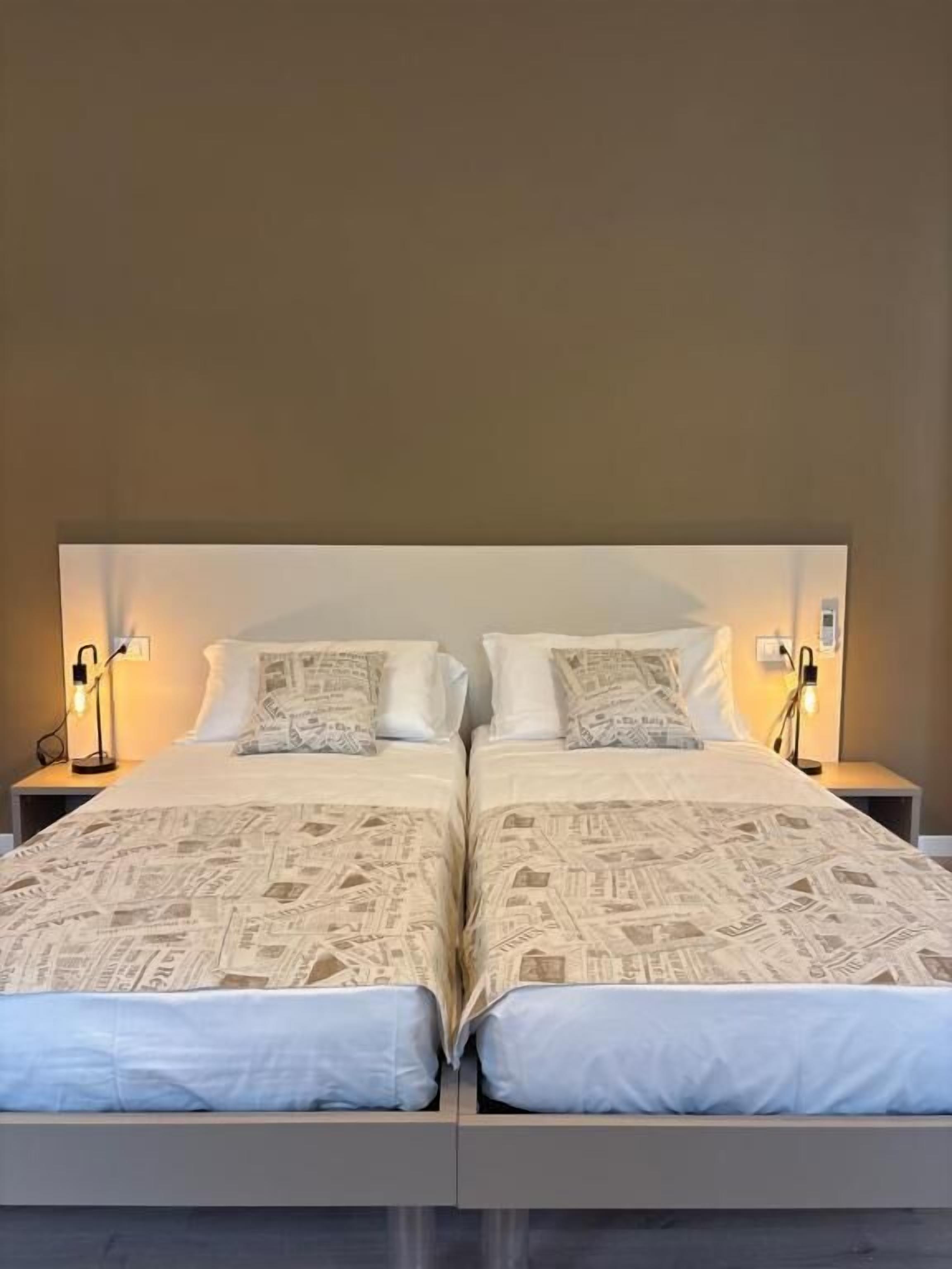 A double bed with a patterned bedspread, two bedside tables with lamps, and a plain wall.