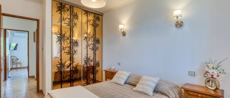 A bedroom with a bed, bedside tables, a wooden nightstand, a decorative wall panel, and a hanging light fixture.