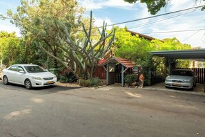 Una calle residencial con dos coches aparcados, una casa pequeña con tejado rojo y un cactus grande al fondo.
