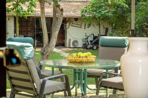 Un patio con una mesa redonda de vidrio, dos sillas, una cesta de mimbre con fruta y un jarrón blanco.