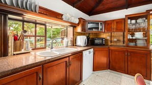 A kitchen with wooden cabinets, a microwave, a sink, and a window overlooking a garden. - Relax in a Garden Oasis by Sea N' Rent (Nahariya)