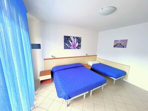 Ett rum med två sängar, ett litet sängbord, en stol, ett fönster med gardiner och en väggmonterad lampa.