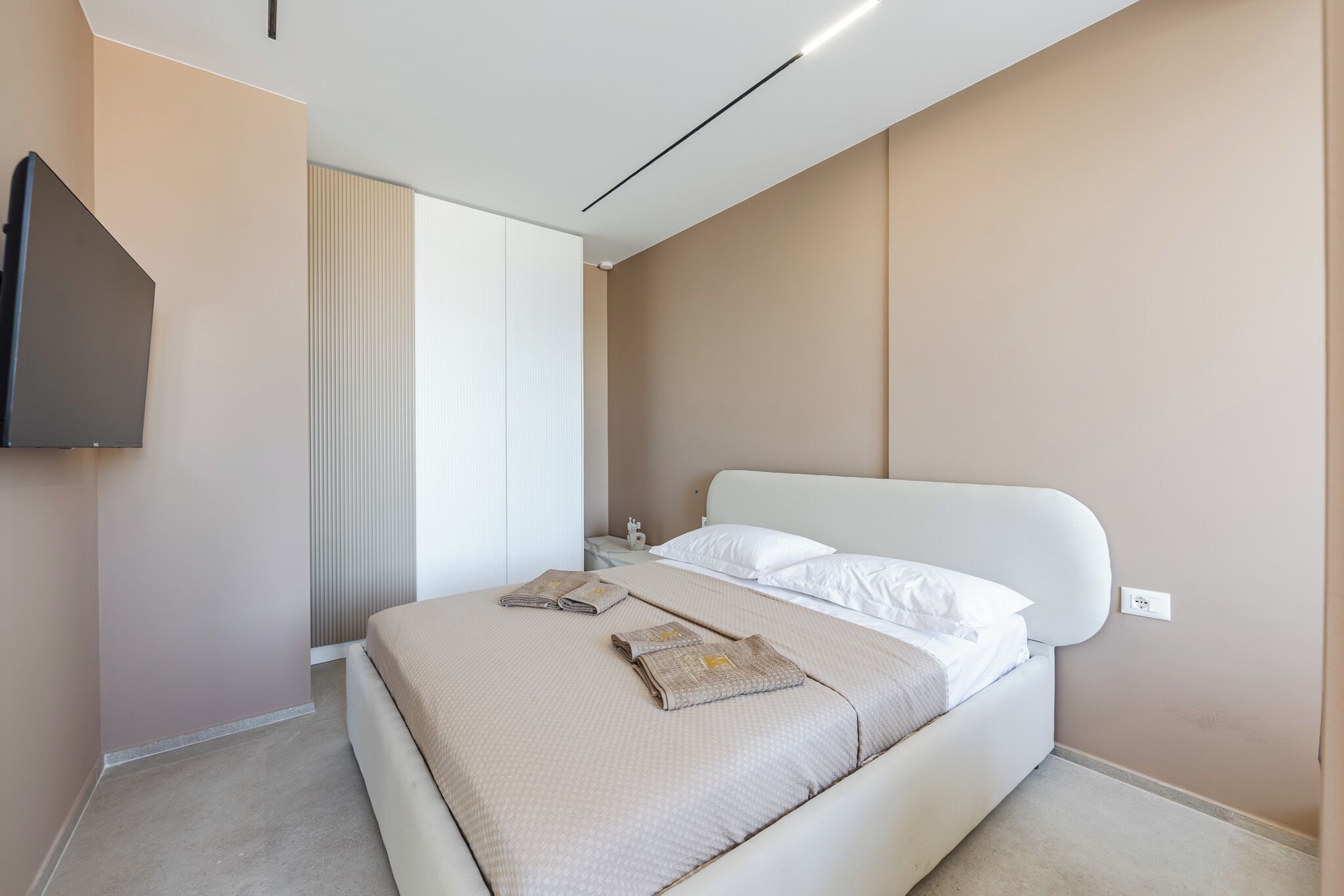 A modern hotel room with a large bed, a flat-screen TV mounted on the wall, and a window with blinds.